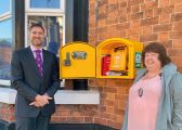 Image of yellow defibrillator box with Stewart Bailey a white man with a beard in a dark suit standing next to Councillor Dawn Clark a white woman in a pink top and grey coat