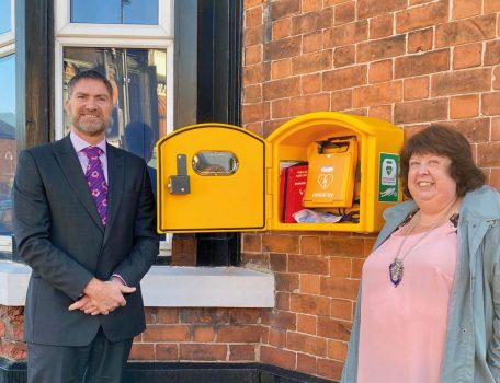 Image of yellow defibrillator box with Stewart Bailey a white man with a beard in a dark suit standing next to Councillor Dawn Clark a white woman in a pink top and grey coat