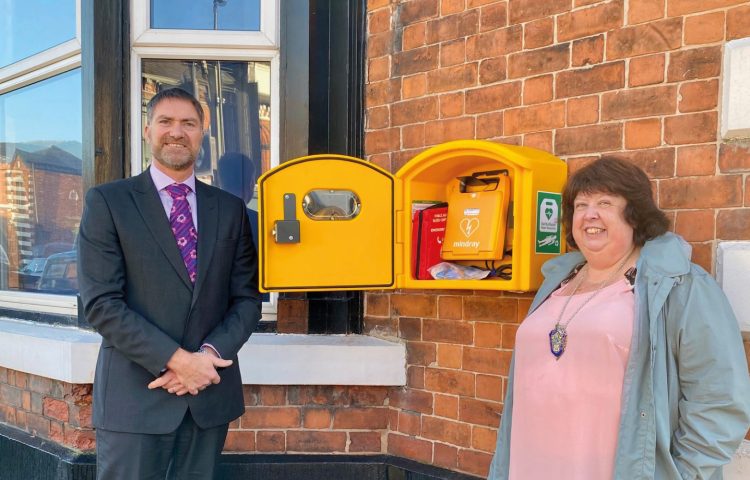 Image of yellow defibrillator box with Stewart Bailey a white man with a beard in a dark suit standing next to Councillor Dawn Clark a white woman in a pink top and grey coat