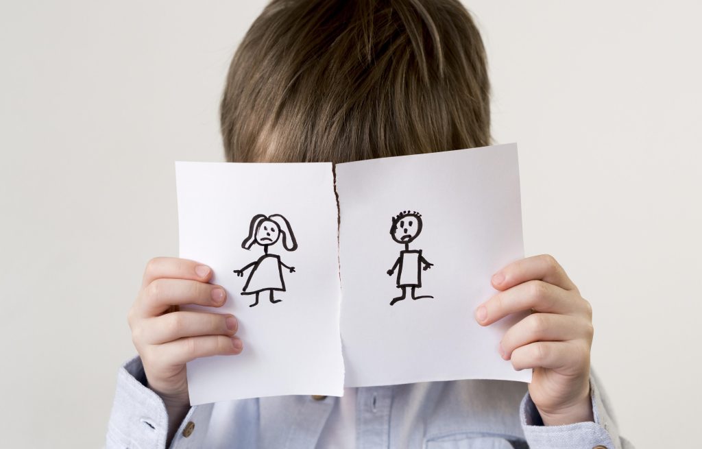 Image shows a young boy with dark hair and a blue shirt with his face covered by a piece of paper torn in half with a stick drawing of a woman and man on each half for the Family Justice Council Blog