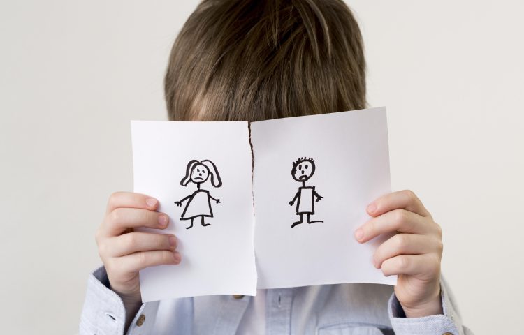 Image shows a young boy with dark hair and a blue shirt with his face covered by a piece of paper torn in half with a stick drawing of a woman and man on each half for the Family Justice Council Blog