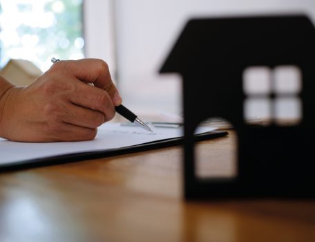 Image of a hand holding a pen and a cardboard cutout of the front of a house sitting on a desk for the Renters Rights Bill