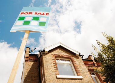 Image of a brick house with upstairs window and a for sale sign outside to represent the mortgage guarantee blog