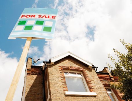 Image of a brick house with upstairs window and a for sale sign outside to represent the mortgage guarantee blog