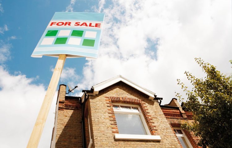 Image of a brick house with upstairs window and a for sale sign outside to represent the mortgage guarantee blog