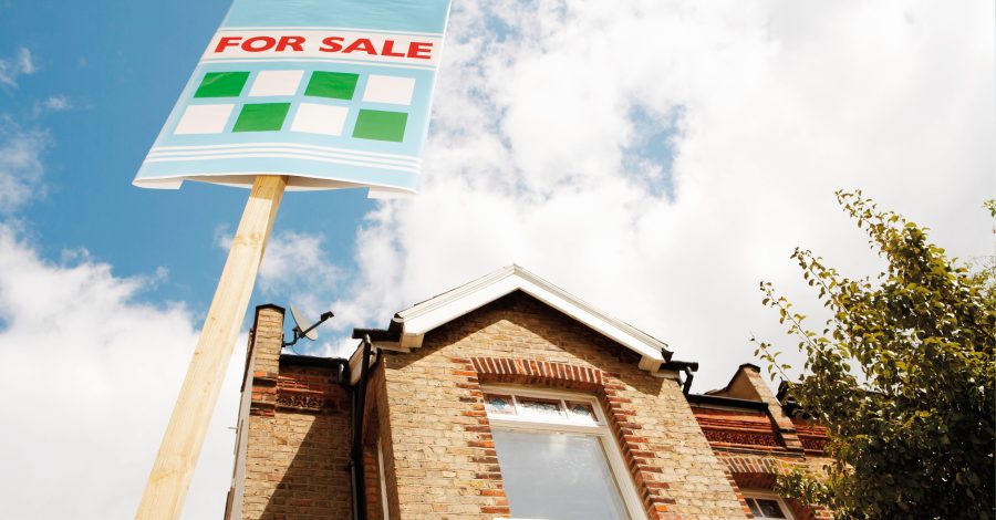 Image of a brick house with upstairs window and a for sale sign outside to represent the mortgage guarantee blog